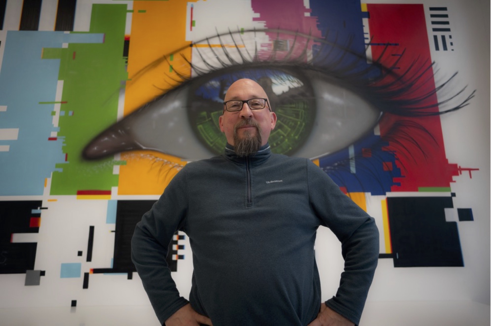 Photo of Alex Counsell, a bearded man in glasses, standing confidently in front of wall art of a giant eye.