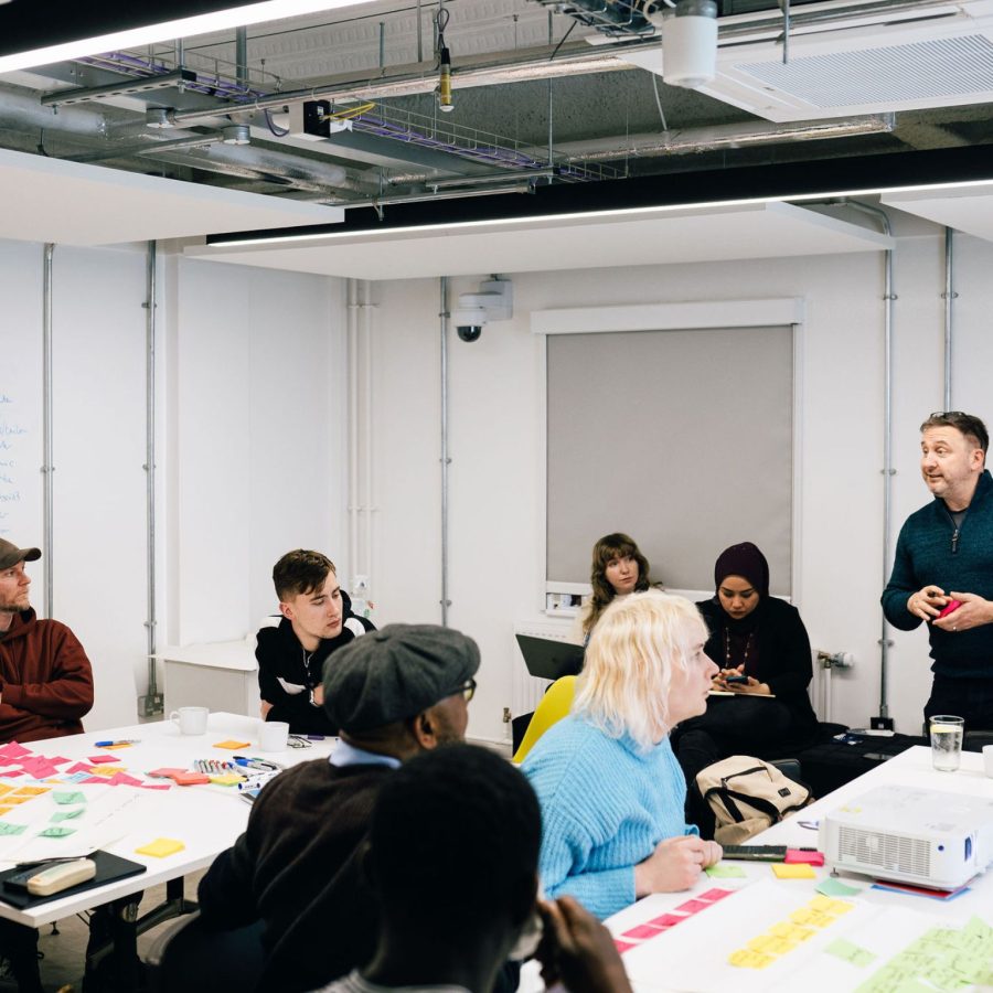People sat around tables with post-it notes during a workshop while a facilitator stands and talks to them.