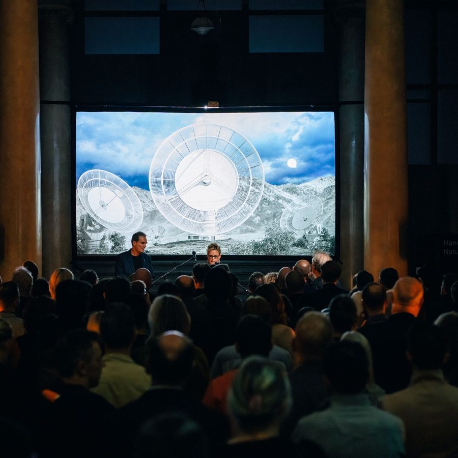 A darkened room with an audience looking at a male and a female speaker in front of a screen showing projected image of transmission dishes.