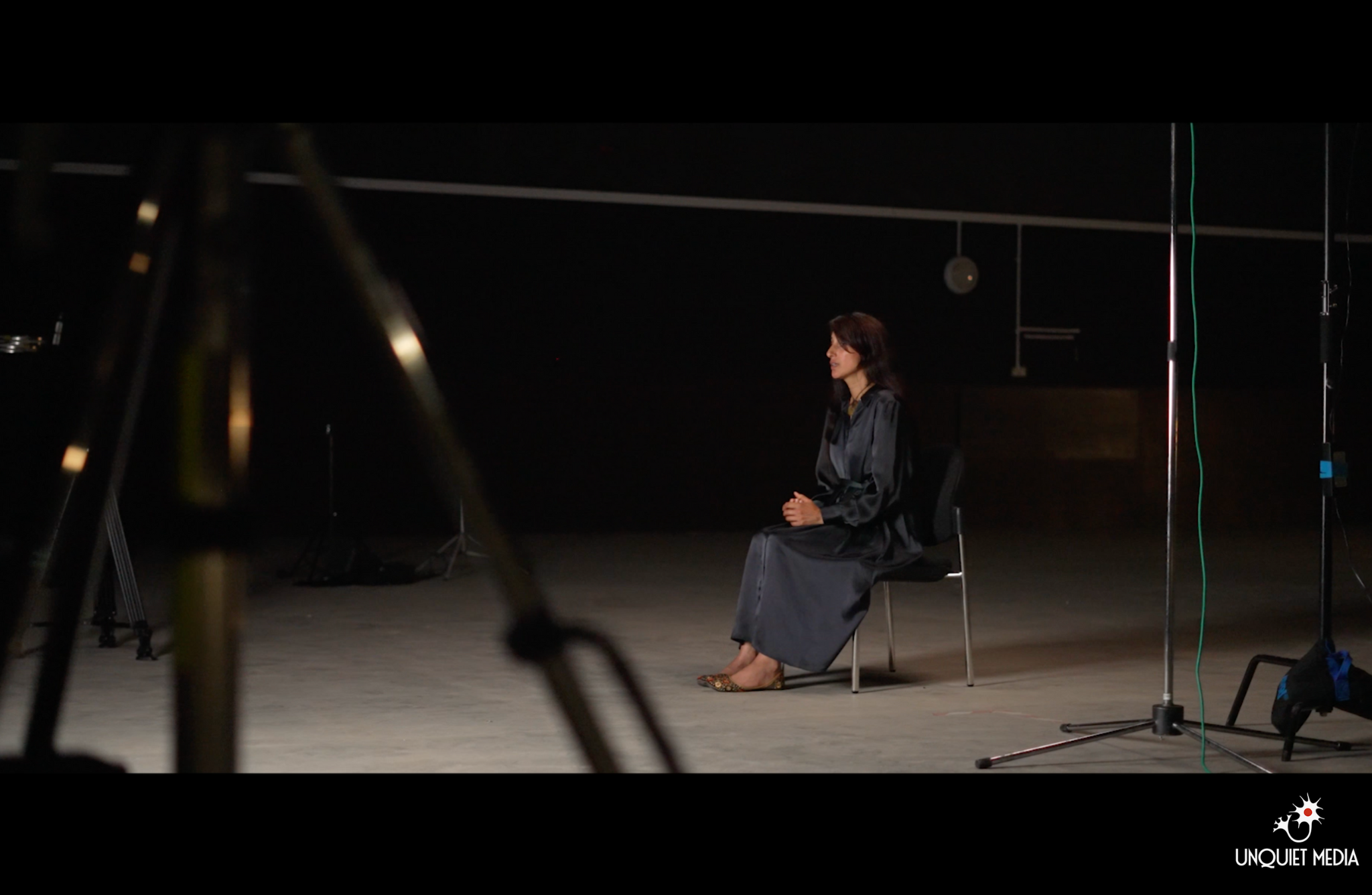 Image Unquiet Media One person is sitting on a black chair in a dimly lit space. There is filming equipment around them. Logo of