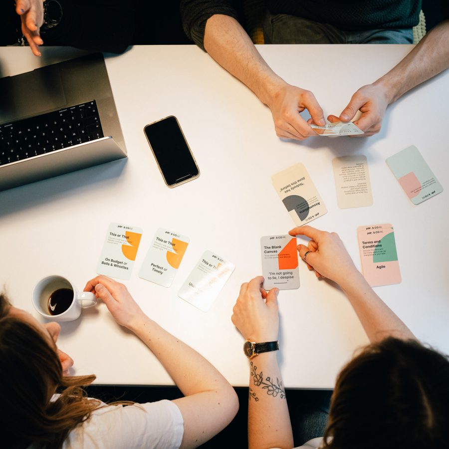 A view from above of a desk where we see the hands of people who are using the core cards together, along with their coffee cups, laptop and smartphone.